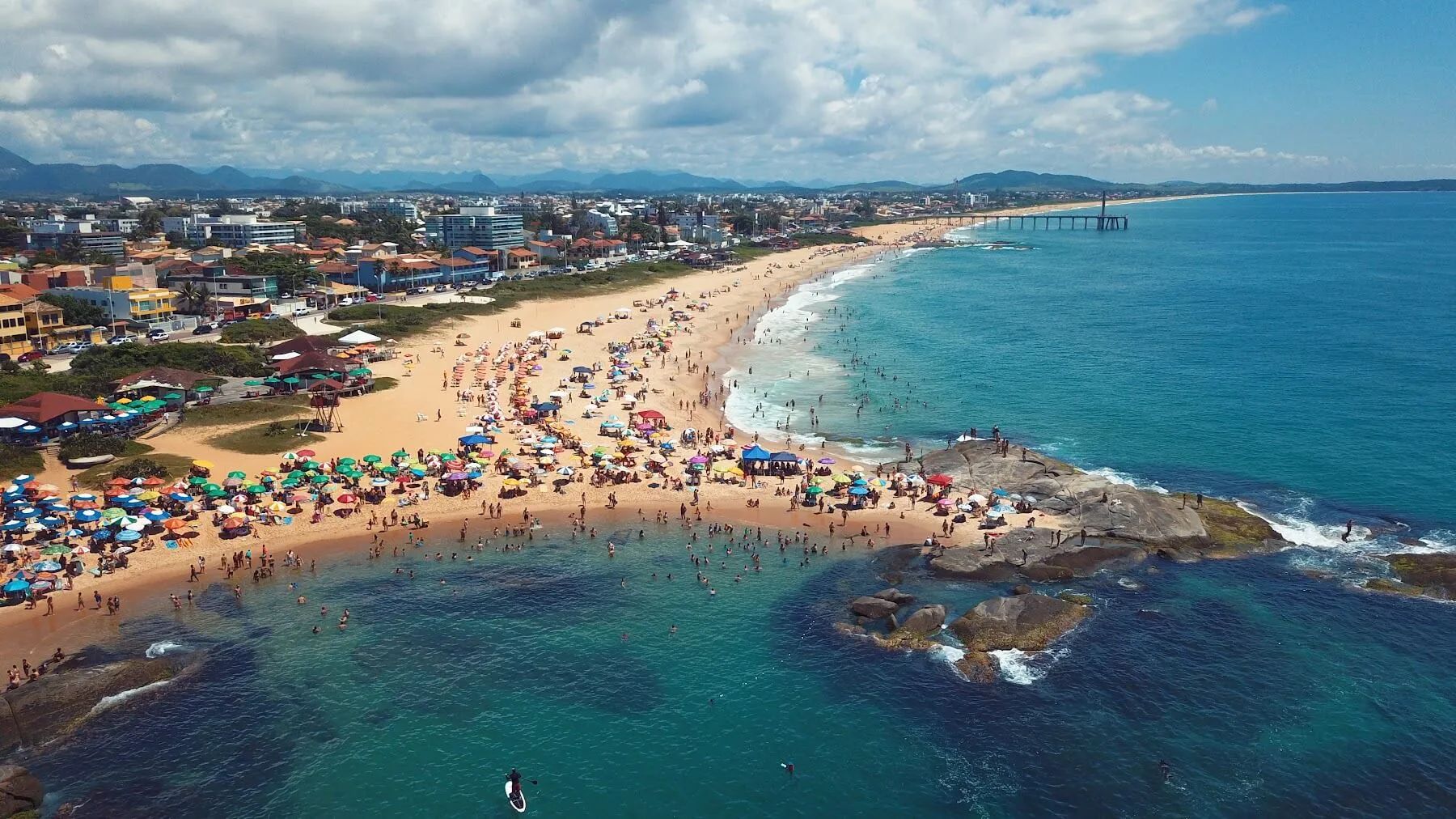 Vista aérea de la playa de Rio das Ostras con sombrillas y muelle al fondo.